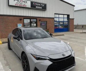 A front three-quarter view of a grey luxury sedan parked inside a brightly lit detailing studio, highlighting dark window tinting and a sleek, high-gloss finish.